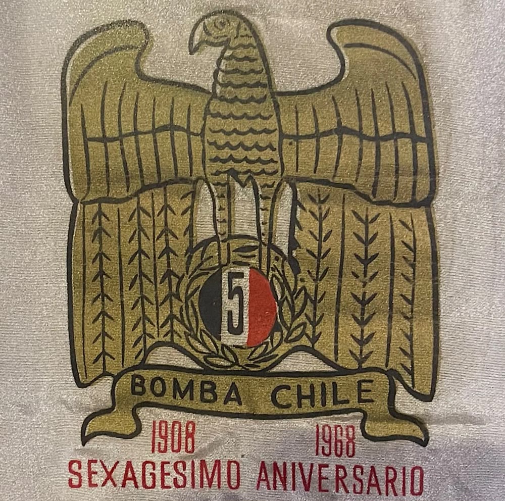 Emblema original del sexagésimo aniversario, impreso en dorado, negro y rojo sobre tela blanca: águila con el 5, cinta Bomba Chile y la leyenda 1908, 1968, Sexagésimo Aniversario